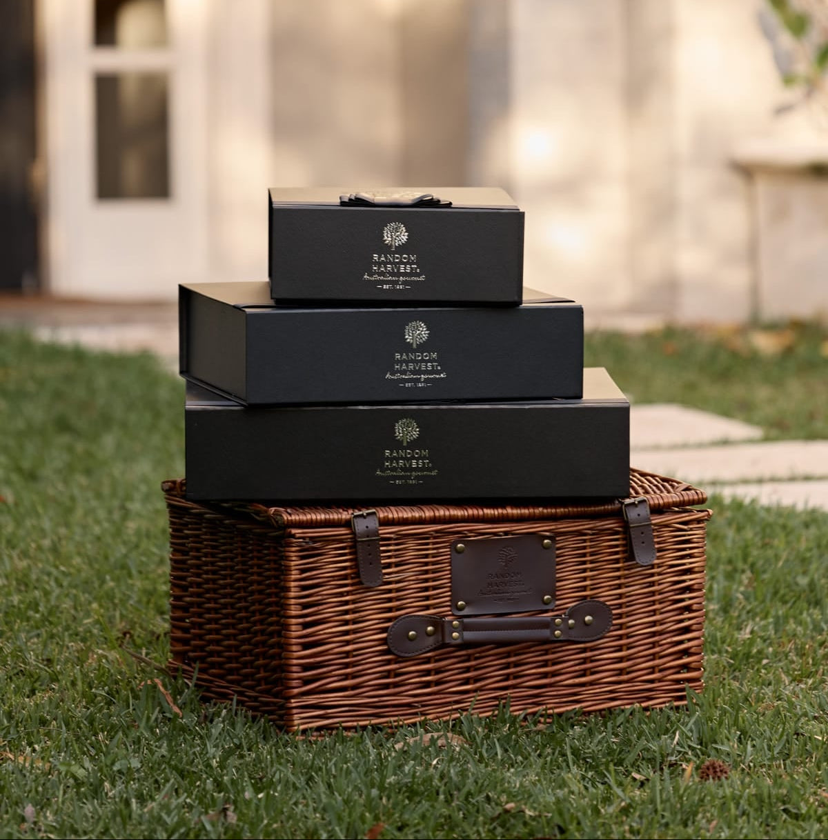 Three stacked black rectangular gift hampers, placed on top of a brown wicker picnic basket on a grassy lawn in front of a softly blurred home facade.